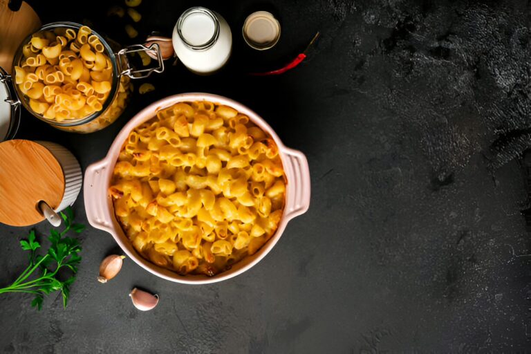 A creamy pasta dish served in a pink casserole dish, with a jar of uncooked pasta, a bottle of milk, and some garlic on a dark surface.