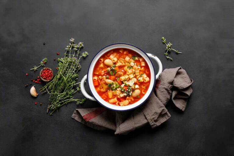 A bowl of colorful vegetable soup with pasta, surrounded by herbs and spices on a dark surface.