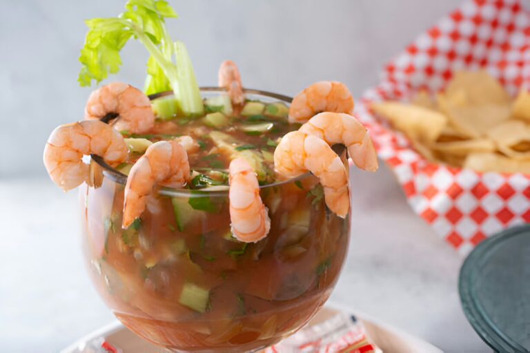 A vibrant shrimp cocktail served in a clear glass, garnished with celery and shrimp, with tortilla chips in the background.
