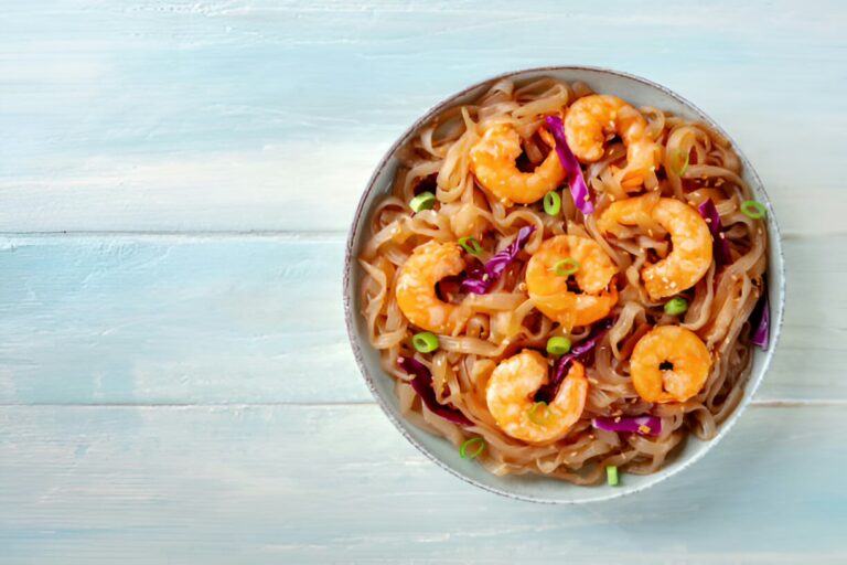 A bowl of shrimp noodles garnished with scallions and purple cabbage on a light blue wooden surface.